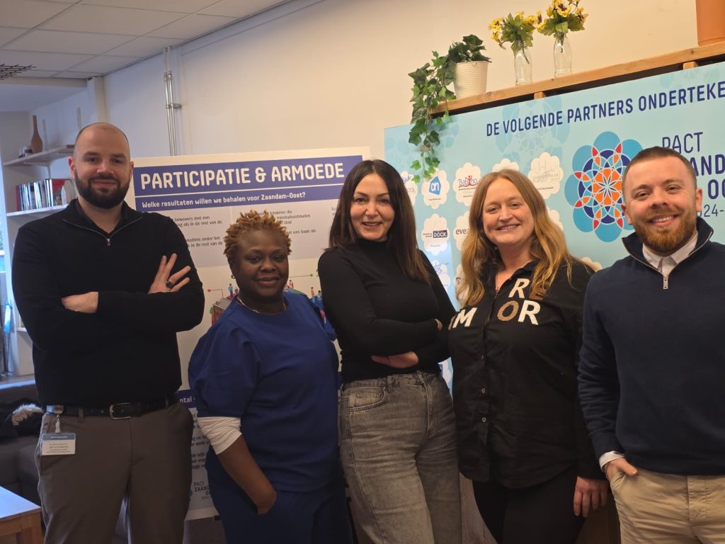 een groep van vijf volwassen poseert voor een bord met participatie en armoede